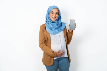 Téléchargez les photos : Sourire femme musulmane enceinte portant hijab Tenez téléphone mobile avec écran vide vide - en image libre de droit