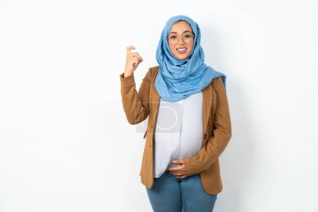 Foto de Mujer musulmana embarazada con hiyab apuntando hacia arriba con la mano mostrando hasta siete dedos gesto en chino lenguaje de signos Q. - Imagen libre de derechos
