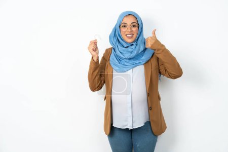 Téléchargez les photos : Femme musulmane enceinte portant le hijab tenant un aligneur d'appareil dentaire invisible et levant le pouce vers le haut, recommandant ce nouveau traitement. Concept de soins dentaires. - en image libre de droit