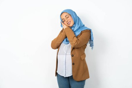 Foto de Hermosa mujer musulmana embarazada con hiyab se apoya en las palmas prensadas cierra los ojos y tiene agradables sueños sonrisa sobre algo - Imagen libre de derechos