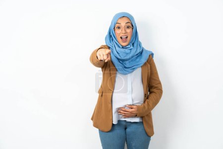 Foto de Emocionado positiva hermosa mujer musulmana embarazada con el dedo índice hijab puntos directamente en usted, ve algo muy divertido. Wow, increíble - Imagen libre de derechos