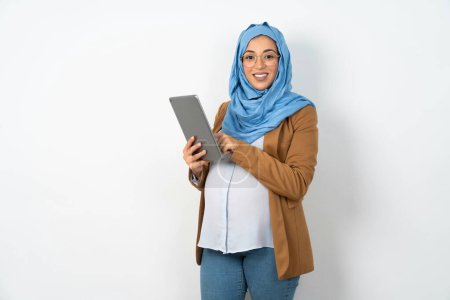 Téléchargez les photos : Photo de heureux gai intelligent belle femme musulmane enceinte portant hijab tenir tablette navigation sur Internet - en image libre de droit