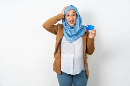 Téléchargez les photos : Impressionné belle femme musulmane enceinte portant hijab avec la main sur la tête tenant la carte bancaire - en image libre de droit
