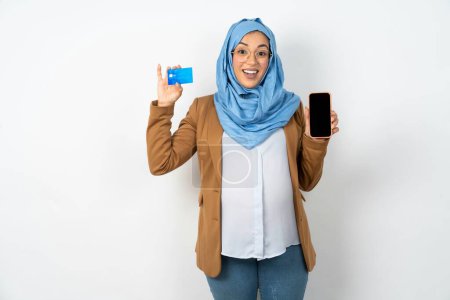 Téléchargez les photos : Belle femme musulmane enceinte portant hijab ouvert compte bancaire, tenant smartphone et carte de crédit, souriant, recommander l'utilisation de l'application de magasinage en ligne - en image libre de droit
