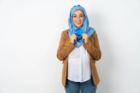 Téléchargez les photos : Photo de belle femme musulmane enceinte portant hijab sourire positif tenir salaire de revenu par carte de crédit - en image libre de droit