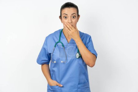 Téléchargez les photos : Médecin arabe émotionnel femme portant des halètements uniformes bleus de l'étonnement, couvre la bouche ouverte avec la paume, semble choqué à la caméra. - en image libre de droit