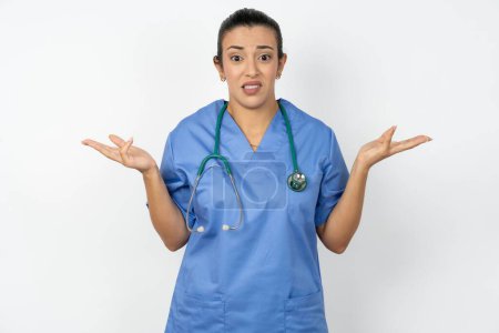 Téléchargez les photos : Médecin arabe sans cervelle femme portant un uniforme bleu haussant les épaules avec hésitation, fait face à une situation douteuse, écarte les paumes, Décision difficile - en image libre de droit
