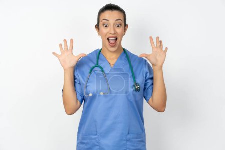 Téléchargez les photos : Optimiste médecin arabe femme portant un uniforme bleu soulève les paumes de joie, heureux de recevoir un cadeau génial de quelqu'un, crie bruyamment, - en image libre de droit