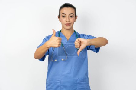 Foto de Árabe médico mujer vistiendo azul uniforme mostrando pulgares hacia arriba y pulgares hacia abajo, difícil elegir concepto - Imagen libre de derechos