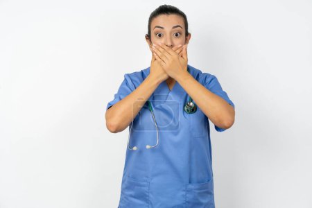 Foto de Árabe médico mujer vistiendo azul uniforme impactado cubriendo la boca con las manos por error. Concepto secreto. - Imagen libre de derechos