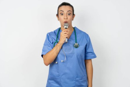 Photo for Very hungry arab doctor woman wearing blue uniform holding spoon into mouth dream of tasty meal - Royalty Free Image