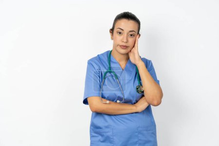 Téléchargez les photos : Médecin arabe très ennuyé femme portant un uniforme bleu tenant la main sur la joue tout en le soutenant avec une autre main croisée, l'air fatigué et malade, - en image libre de droit