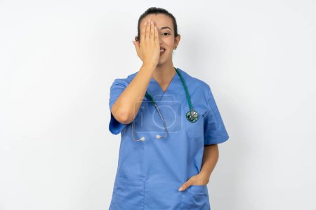 Téléchargez les photos : Médecin arabe femme portant un uniforme bleu couvrant un oeil avec sa main, sourire confiant sur le visage et l'émotion surprise. - en image libre de droit