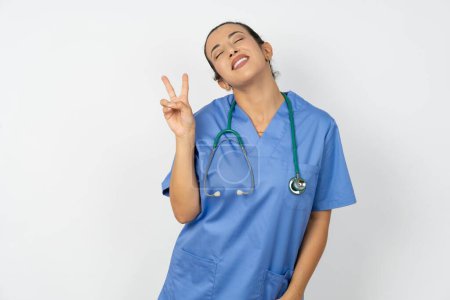Foto de Árabe médico mujer vistiendo uniforme azul sonriendo con la cara feliz guiño a la cámara haciendo signo de victoria. Número dos.. - Imagen libre de derechos