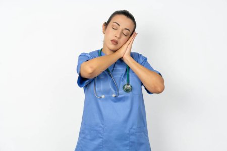 Téléchargez les photos : Arabe médecin femme portant uniforme bleu dormir fatigué rêver et posant avec les mains ensemble tout en souriant avec les yeux fermés. - en image libre de droit