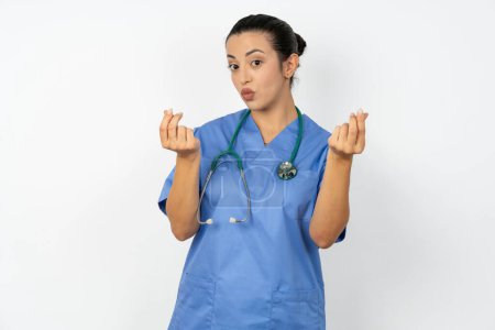 Téléchargez les photos : Médecin arabe femme portant un uniforme bleu faisant geste d'argent avec les mains, demandant le paiement du salaire, entreprise millionnaire - en image libre de droit