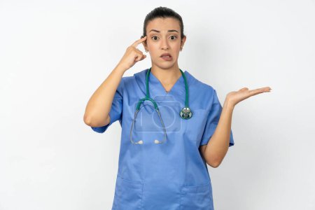 Téléchargez les photos : Médecin arabe femme portant uniforme bleu confus et agacé avec la paume ouverte montrant l'espace de copie et pointant du doigt vers le front. Penses-y.. - en image libre de droit