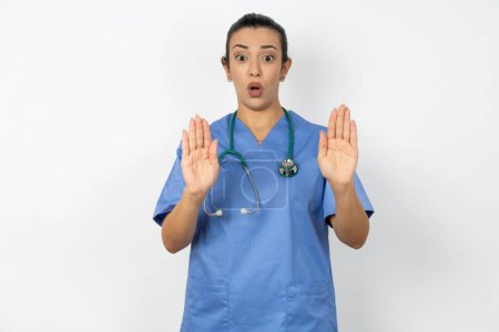 Foto de Médico árabe mujer vistiendo uniforme azul Manos alejadas palmas que muestran rechazo y negación con miedo y expresión repugnante. Detente y prohibido. - Imagen libre de derechos