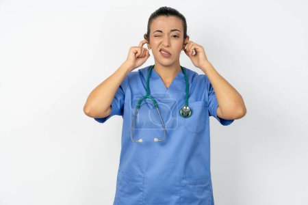 Téléchargez les photos : Médecin arabe femme portant uniforme bleu couvrant les oreilles avec les doigts avec une expression agacée pour le bruit de la musique forte. Concept sourd. - en image libre de droit