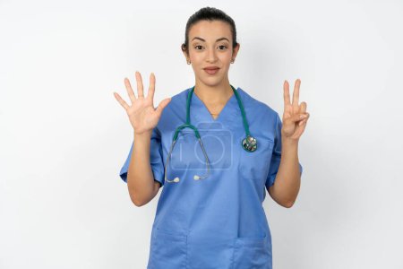 Téléchargez les photos : Arabe médecin femme portant uniforme bleu montrant et pointant vers le haut avec les doigts numéro sept tout en souriant confiant et heureux. - en image libre de droit