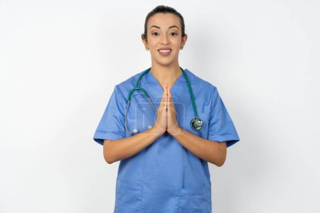 Foto de Árabe médico mujer vistiendo azul uniforme orando con las manos juntas pidiendo perdón sonriendo confiado. - Imagen libre de derechos