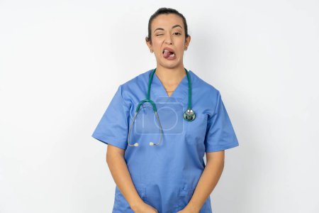 Téléchargez les photos : Médecin arabe femme portant uniforme bleu langue collante heureux avec une expression drôle. Concept d'émotion. - en image libre de droit
