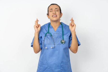 Foto de Árabe médico mujer vistiendo azul uniforme gesto dedos cruzados sonriendo con esperanza y los ojos cerrados. Suerte y concepto supersticioso. - Imagen libre de derechos