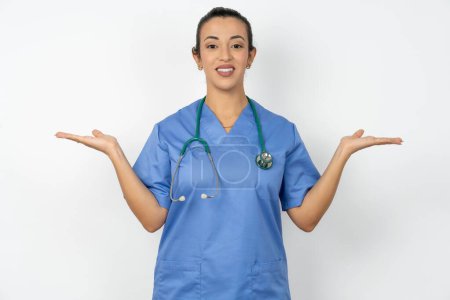 Photo for Cheerful cheery optimistic arab doctor woman wearing blue uniform  holding two palms copy space - Royalty Free Image