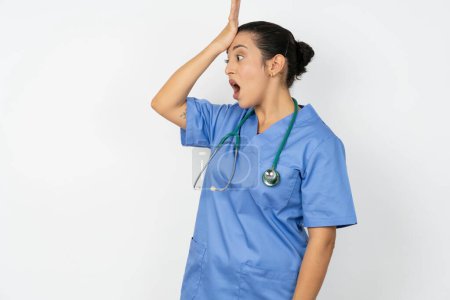 Téléchargez les photos : Médecin arabe femme portant un uniforme bleu surpris avec la main sur la tête pour erreur, rappelez-vous erreur. Oublié, mauvais concept de mémoire. - en image libre de droit