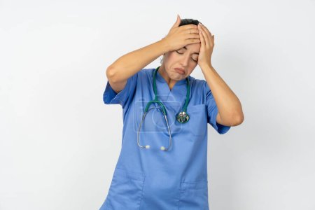Foto de Árabe médico mujer vistiendo uniforme azul que sufre de fuerte dolor de cabeza desesperado y estresado debido al exceso de trabajo. Concepto de depresión y dolor. - Imagen libre de derechos