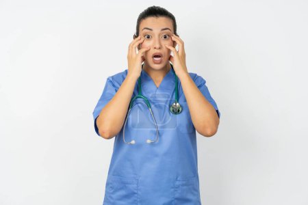 Téléchargez les photos : Stupéfiée médecin arabe femme portant un uniforme bleu exprime l'excitation et le frisson, garde la mâchoire tombée, mains sur les joues, a les yeux sortis - en image libre de droit