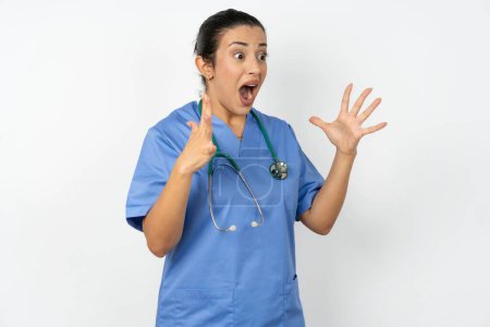 Foto de Árabe médico mujer vistiendo azul uniforme grita fuerte, mantiene los ojos abiertos y las manos tensas. - Imagen libre de derechos