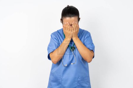 Téléchargez les photos : Médecin arabe femme portant un uniforme bleu couvrant son visage avec ses mains, étant dévastée et pleurant. Triste concept - en image libre de droit