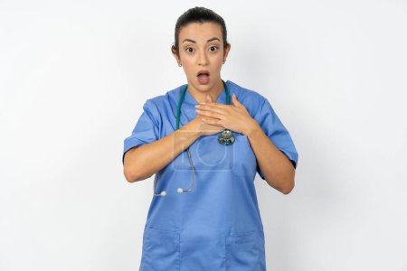 Téléchargez les photos : Médecin arabe effrayé femme portant un uniforme bleu regarde avec une expression effrayée, garde les mains sur la poitrine, étant perplexe de remarquer quelque chose d'étrange, Les gens, réaction silencieuse et émotions. - en image libre de droit