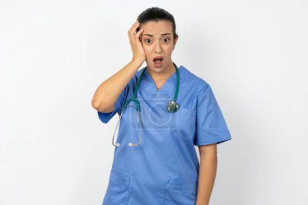 Téléchargez les photos : Embarrassé médecin arabe femme portant un uniforme bleu avec une expression choquée, exprime une grande stupéfaction, modèle perplexe pose à l'intérieur - en image libre de droit