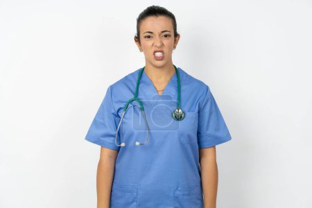 Photo for Arab doctor woman wearing blue uniform keeps teeth clenched, frowns face in dissatisfaction, irritated because of much duties. - Royalty Free Image