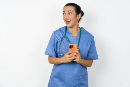 Photo for Arab doctor woman wearing blue uniform holding a smartphone and looking sideways at blank copyspace. - Royalty Free Image