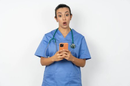 Téléchargez les photos : Choqué arabe médecin femme portant uniforme bleu ouvre la bouche tenir téléphone lecture annonce incroyable gros prix - en image libre de droit