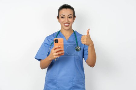Téléchargez les photos : Portrait de médecin arabe femme portant uniforme bleu en utilisant et textos avec smartphone heureux avec un grand sourire faire signe ok, pouce levé avec les doigts, excellent signe - en image libre de droit