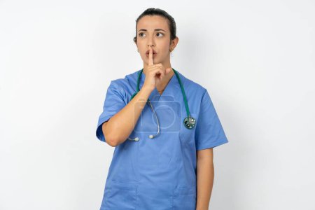 Téléchargez les photos : Femme arabe médecin en uniforme avec stéthoscope geste de silence garde index aux lèvres fait signe de silence. Demande de ne pas partager le secret - en image libre de droit