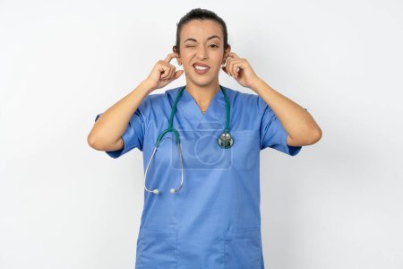 Photo for Happy arab woman doctor in  uniform with stethoscope ignores loud music and plugs ears with fingers asks to turn off sound - Royalty Free Image