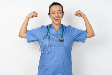 Foto de Mujer árabe fuerte médico en uniforme con estetoscopio con sonrisa dentada, levanta los brazos y muestra bíceps. Mira mis músculos.! - Imagen libre de derechos