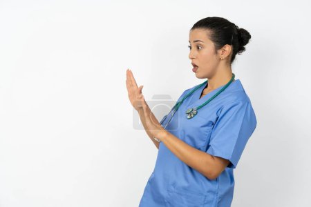 Foto de Mujer árabe disgustado médico en uniforme con estetoscopio mantiene las manos hacia el espacio vacío y pide no acercarse ve algo desagradable - Imagen libre de derechos