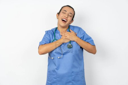Foto de Mujer árabe médico en uniforme con estetoscopio expresa felicidad, ríe agradablemente, mantiene las manos en el corazón - Imagen libre de derechos