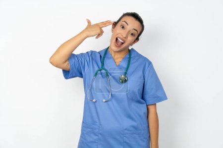 Photo for Arab woman doctor in  uniform with stethoscope  foolishness around shoots in temple with fingers makes suicide gesture. Funny model makes finger gun pistol - Royalty Free Image