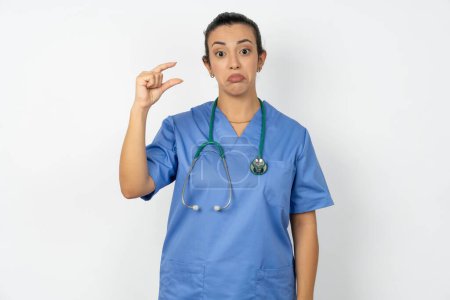 Téléchargez les photos : Femme arabe médecin en uniforme avec porte-monnaie stéthoscope lèvre et gestes à la main, montre quelque chose de très peu. - en image libre de droit