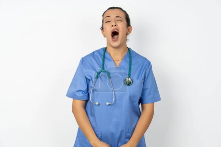 Téléchargez les photos : Femme arabe médecin en uniforme avec des bâillements stéthoscopiques avec des stands à bouche ouverte. routine quotidienne du matin - en image libre de droit
