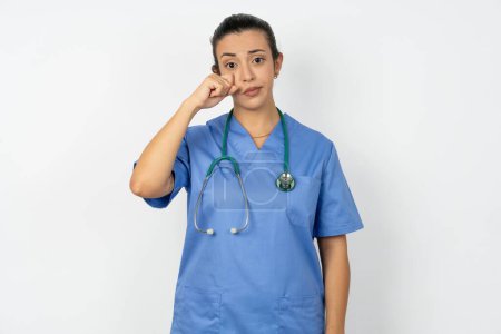 Foto de Decepcionado mujer árabe abatido médico en uniforme con estetoscopio limpia lágrimas se encuentra estresado con expresión sombría. Emoción negativa - Imagen libre de derechos