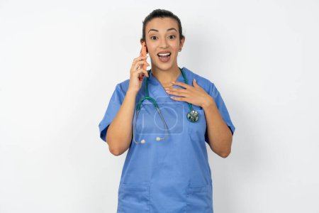 Foto de Mujer árabe sonriente médico vistiendo azul uniforme conversaciones a través del teléfono celular, disfruta de una agradable gran conversación. Personas, tecnología, concepto de comunicación - Imagen libre de derechos