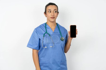 Téléchargez les photos : Médecin arabe femme portant uniforme bleu tient un nouveau téléphone portable et semble mystérieux de côté montre l'affichage vierge de cellulaire moderne - en image libre de droit